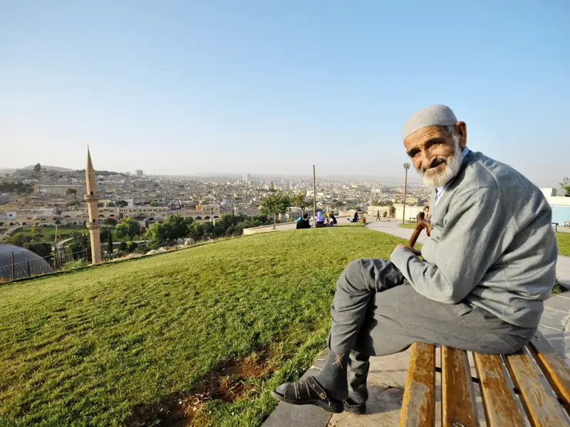 Ein freundlich lächelnder, älterer Mann sitzt auf einer Parkbank mit weitem Blick über die Dächer der türkischen Stadt Sanliurfa.