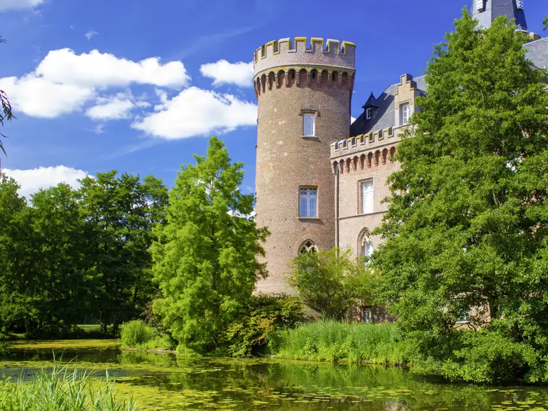 Das Wasserschloss Moyland am Niederrhein mit seinem markanten Backsteinturm, umgeben vom Wassergraben und grünen Bäumen, ein Highlight jeder Rundreise mit Studiosus