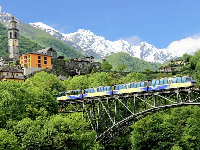 Die Centovalli-Bahn fährt auf einer Studienreise im Tessin über ein Viadukt bei Intragna vor einer Kulisse aus schneebedeckten Alpen.