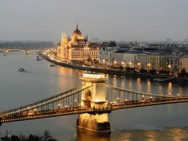 Auf unserer Gruppenreise mit Studiosus genießen wir die abendliche Ansicht der beleuchteten Szechenyi-Kettenbrücke über der Donau und des ungarischen Parlamentsgebäudes in Budapest.