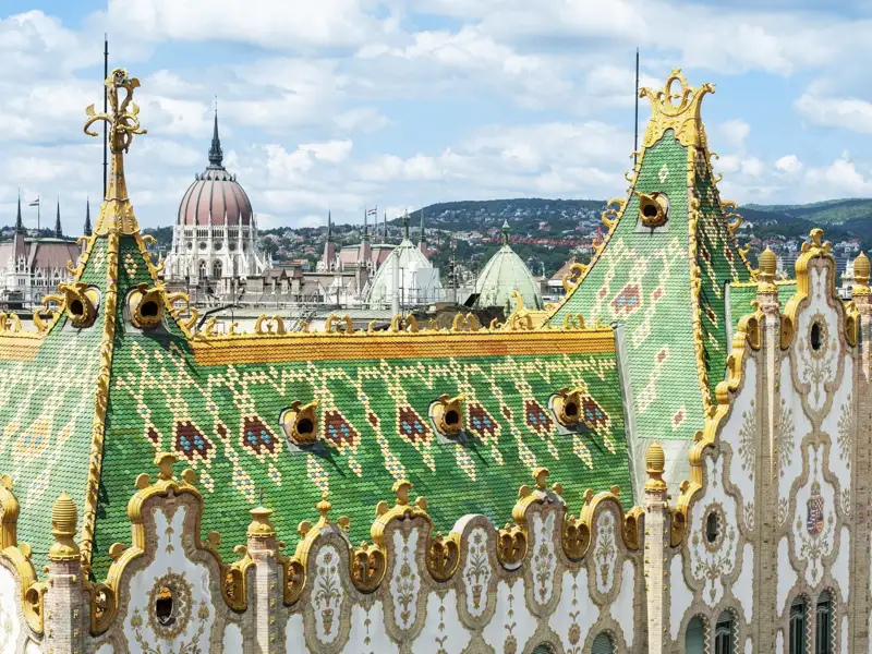 Verziertes grünes Ziegeldach der Postsparkasse im Jugendstil in Budapest, im Hintergrund das ungarische Parlamentsgebäude, ein architektonisches Highlight auf unserer Studiosus Gruppenreise.