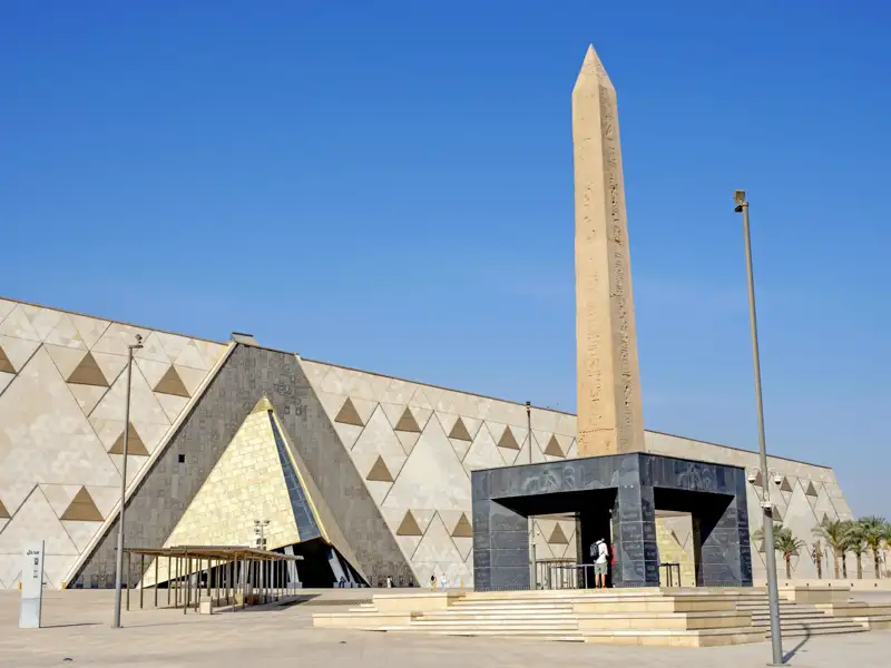 Die moderne Fassade des Grand Egyptian Museum in Gizeh, davor ein hoher, antiker Obelisk unter strahlend blauem Himmel, ein architektonisches Highlight unserer Gruppenreise.