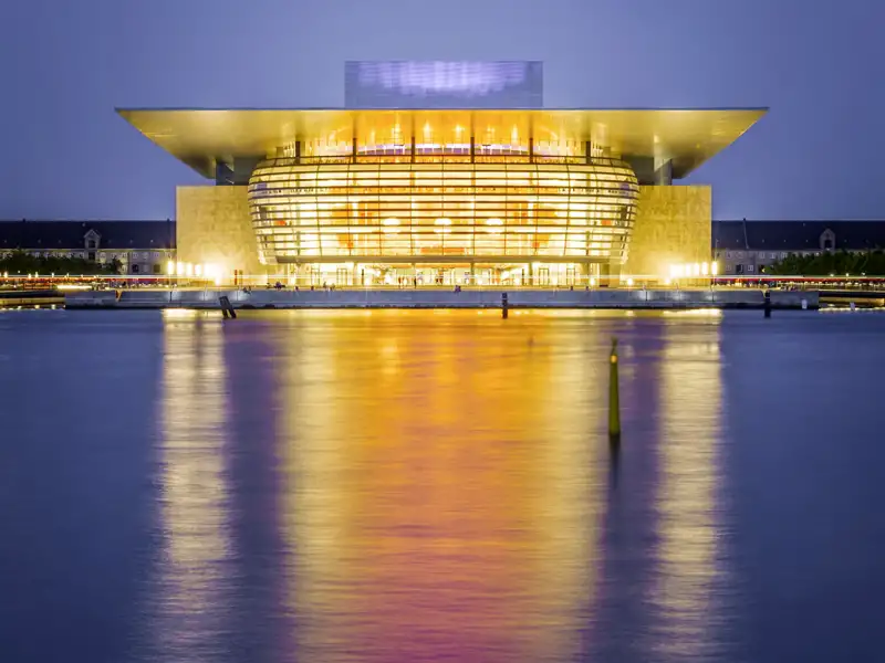 Das beleuchtete Opernhaus Kopenhagen bei Nacht, dessen Lichter sich im Wasser spiegeln.