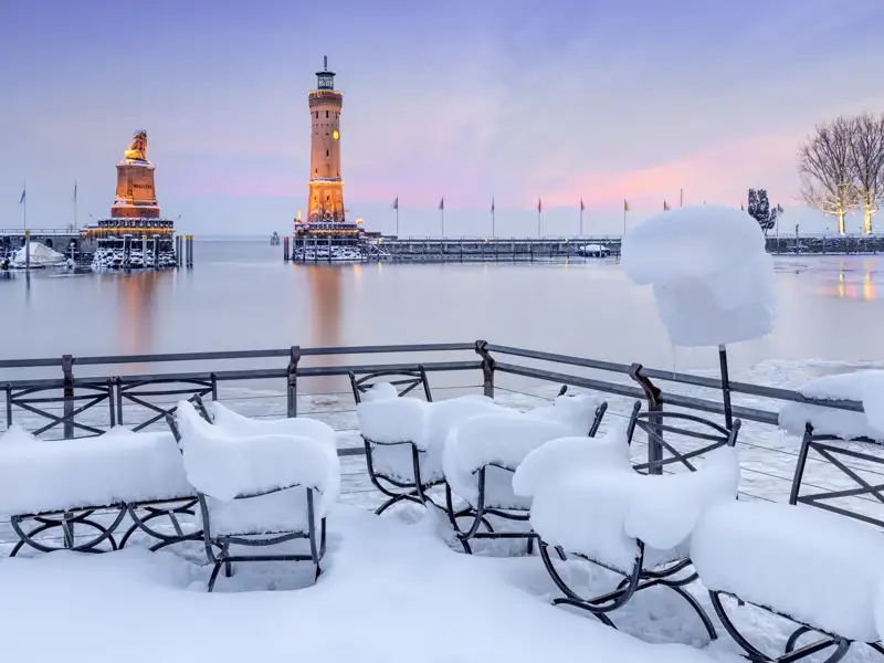 Winterliche Hafeneinfahrt von Lindau am Bodensee mit Bayerischem Löwen und Leuchtturm, im Vordergrund schneebedeckte Stühle.