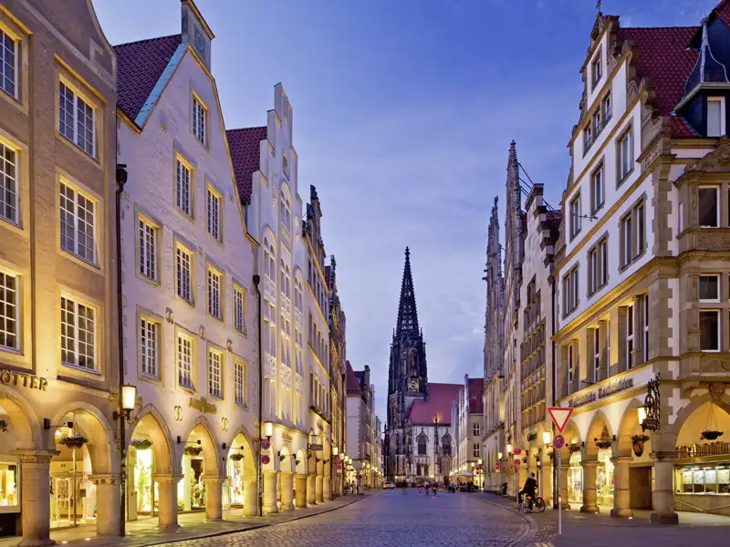 Der Prinzipalmarkt in Münster bei Abenddämmerung: Die beleuchteten Giebelhäuser und die Lambertikirche im Hintergrund schaffen eine magische Kulisse. Ideal für entspanntes Schlendern durch die Altstadt auf dieser Studiosus Gruppenreise