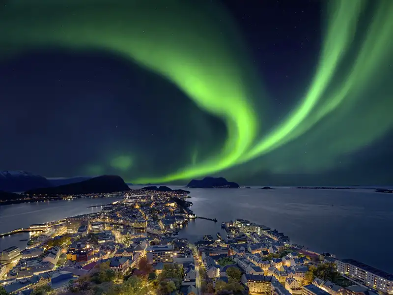 Grüne, tanzende Nordlichter am Sternenhimmel über der beleuchteten Küstenstadt Ålesund und der Fjordlandschaft Norwegens auf ihrer Studiosus Eventreise.