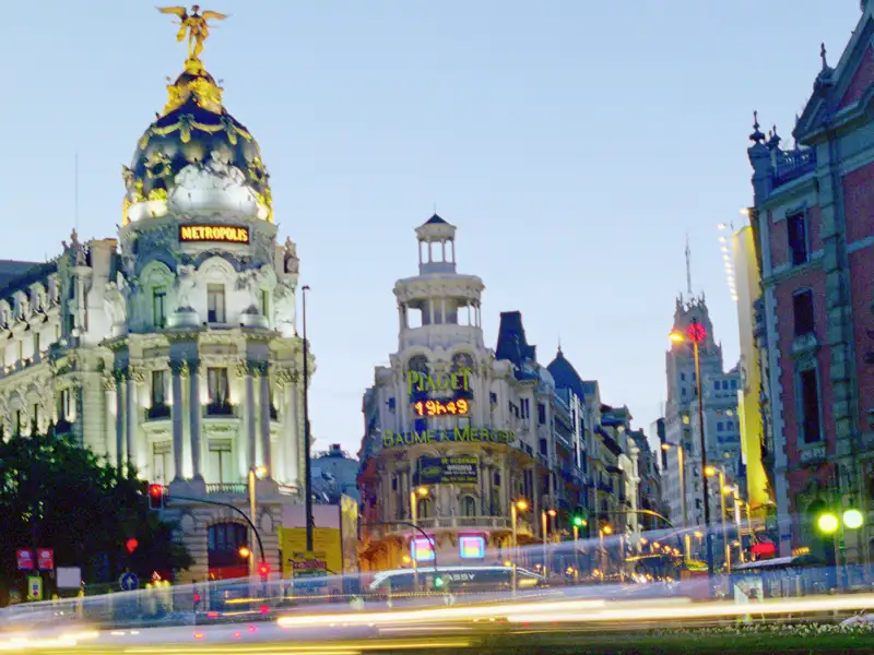 Das ikonische Metropolis-Gebäude an der Prachtstraße Gran Vía in Madrid leuchtet in der Abenddämmerung über Lichtspuren. Ein Highlight Ihrer Studiosus Eventreise.