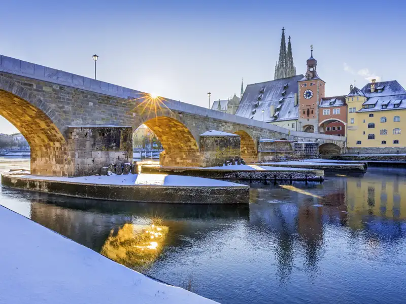 Die Steinerne Brücke über der Donau in Regensburg an einem sonnigen Wintertag mit Schnee und den Domspitzen im Hintergrund. Ein schönes Fotomotiv während Ihrer Studienreise.