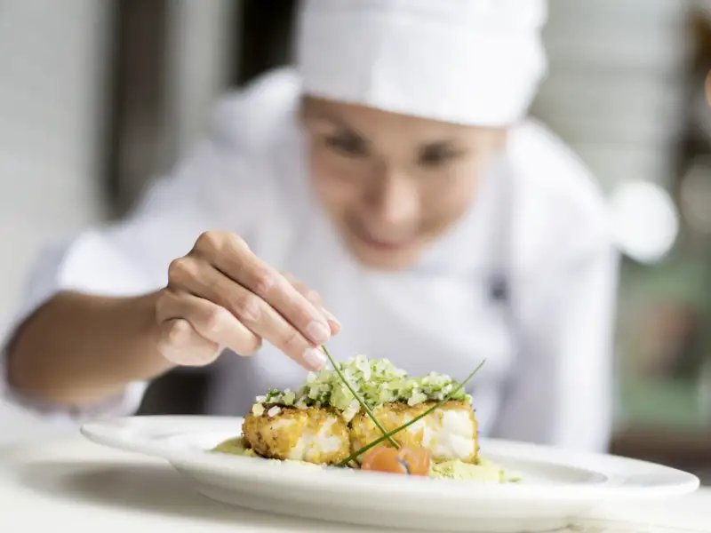 Eine Köchin in weißer Uniform garniert sorgfältig ein exquisites Fischgericht mit frischen Kräutern auf einem weißen Teller.
