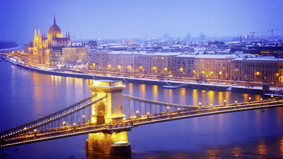 Blick auf die beleuchtete Kettenbrücke über der Donau und das verschneite Parlamentsgebäude in Budapest bei Dämmerung.