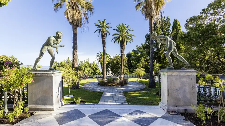 Bronzestatuen im sonnigen Schlossgarten von Sisis Achilleion-Palast auf Korfu, umgeben von hohen Palmen und grünen Pflanzen, ein Highlight unserer Studiosus Gruppenreise.