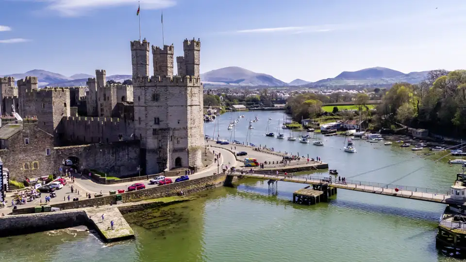 Luftaufnahme des UNESCO-Welterbes Caernarfon Castle am Hafen von Caernarfon mit Segelbooten vor walisischer Bergkulisse, ein Highlight unserer Gruppenreise mit Studiosus
