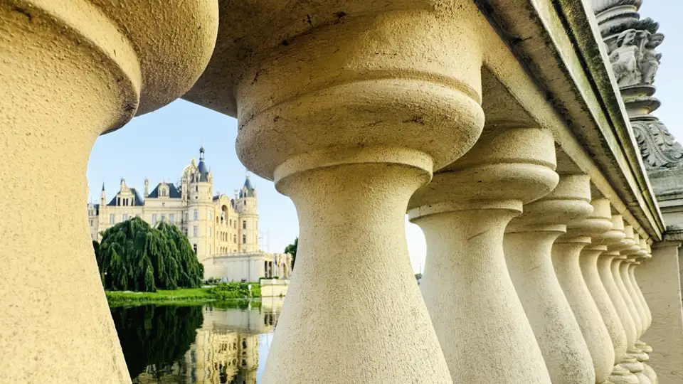 Das verspielte Schweriner Schloss, durch die Baluster einer Steinbrücke gesehen, spiegelt sich im ruhigen Wasser des Sees, ein Highlight unserer Studiosus Gruppenreise an die Ostsee.