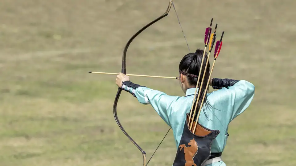 Rückansicht einer Bogenschützin in hellblauer Tracht, die bei den World Nomad Games in Kirgisistan einen Pfeil anlegt, ein Highlight unserer Gruppenreise mit Studiosus.