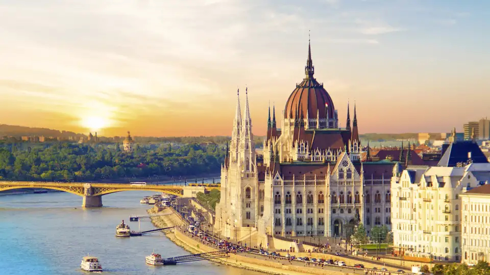 Das ungarische Parlamentsgebäude in Budapest am Donauufer im goldenen Licht des Sonnenuntergangs, Ausflugsboote auf dem Fluss, ein Ausblick, den wir auf einer Studiosus Rundreise genießen.