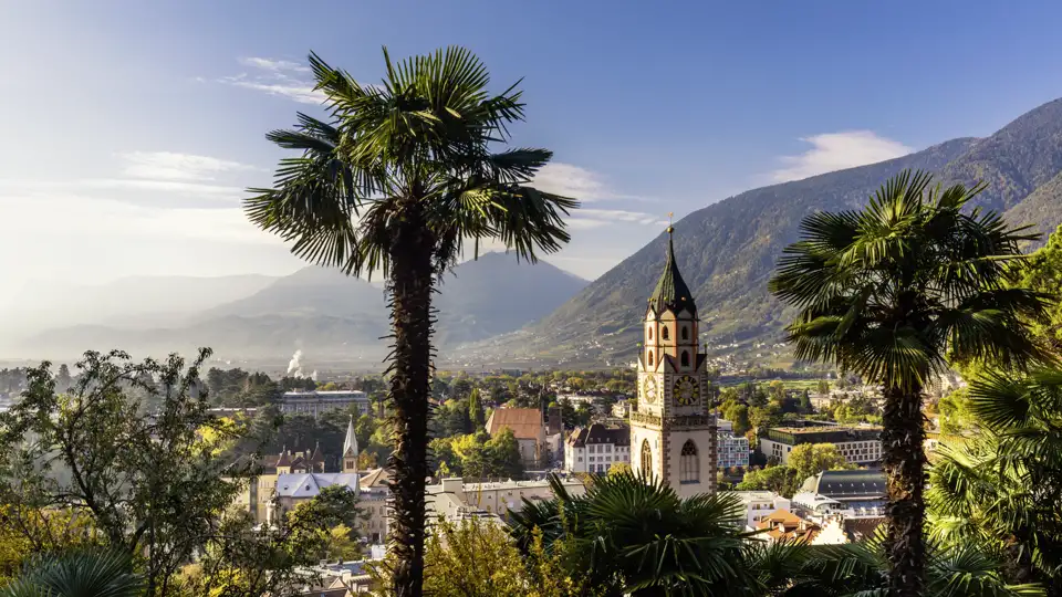 Mediterranes Flair in Meran: Palmen rahmen den Blick auf die Pfarrkirche St. Nikolaus und die Berglandschaft Südtirols, welche Sie auf der Eventreise bestaunen können.