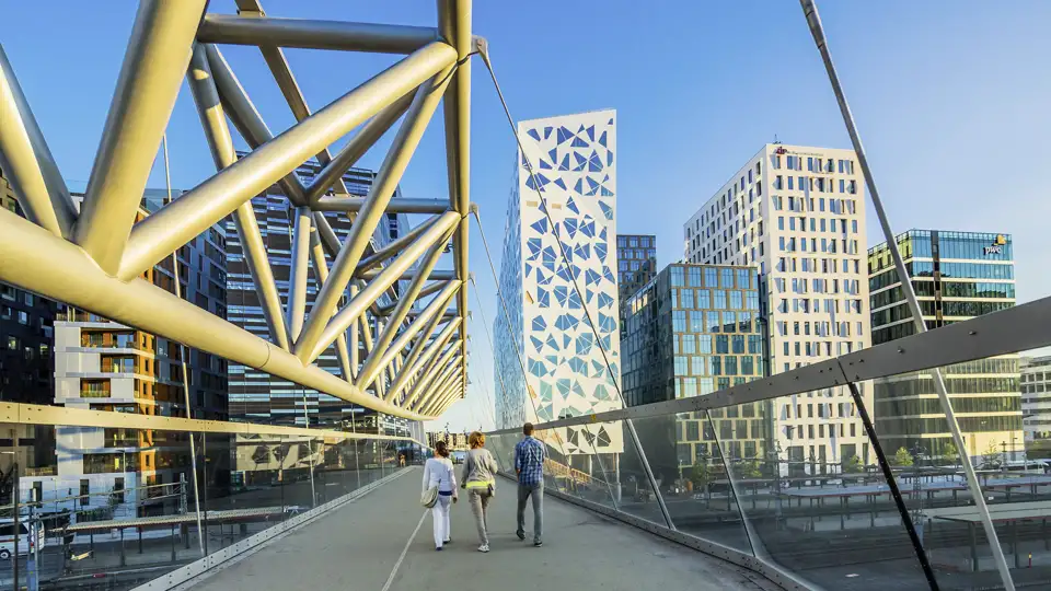 Kleine Reisegruppe auf einer Fußgängerbrücke vor den modernen Hochhäusern des Björvika-Viertels in Oslo bei Sonnenschein.