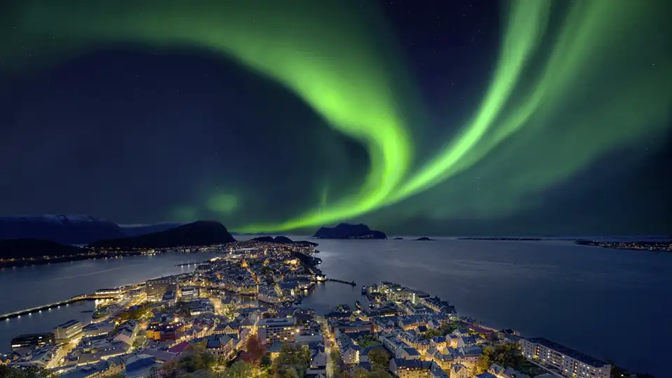 Grüne, tanzende Nordlichter am Sternenhimmel über der beleuchteten Küstenstadt Ålesund und der Fjordlandschaft Norwegens auf ihrer Studiosus Eventreise.