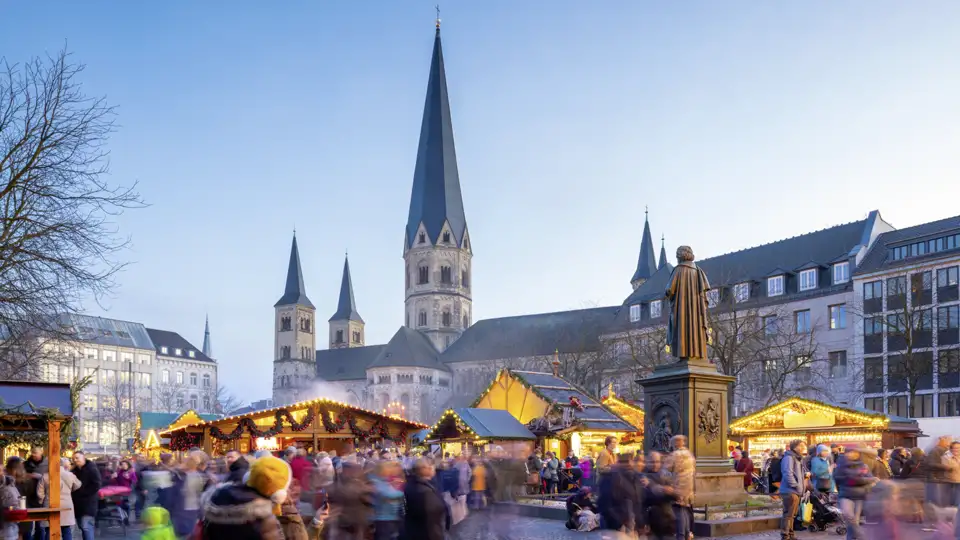 Festlich beleuchteter Weihnachtsmarkt auf dem Bonner Münsterplatz mit dem Beethoven-Denkmal und dem Münster bei Dämmerung. Ein Highlight Ihrer Studiosus Silvesterreise.