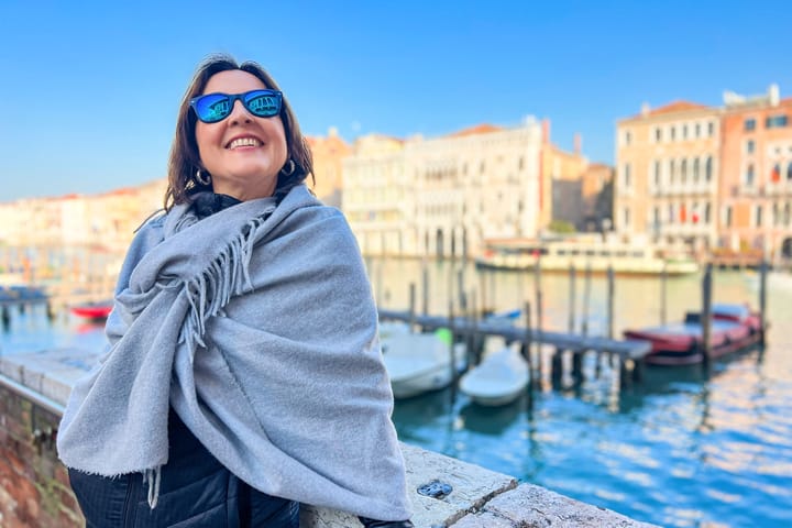 Eine lachende Frau mit Schal um die Schulter steht in Venedig am Wasserrand.